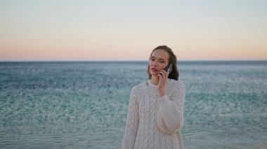 Okyanus ufkunda akıllı telefondan konuşan bir kadın. Gülümseyen kadın gezgin cep telefonunu arıyor. Akşamları sakin deniz dalgalarının yanında duruyor. Mutlu kız gün batımında cep telefonu sohbetinden zevk alıyor..