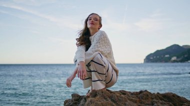 Rocky Beach 'te poz veren kız rüzgarlı yaz akşamının tadını çıkarıyor. Mutlu gezgin kadın sabah deniz manzarası kamerasına bakıyor. Kıyı taşlarına çömelmiş muhteşem bir model. Uzun saçlı, dinlenen bir kadın.