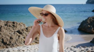 Güneşli sahilde hasır şapka takıp pina colada içen bir kız. Deniz ufkunun önünde kumu yudumlayan güzel bir model. Gülümseyen kadın güneş gözlüğünü çıkarıp kameraya bakıyor. Yaz tatili