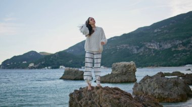 Kadınlar kıyı kayalıklarında rüzgarlı deniz sabahlarını severler. Kaygısız, rahat bir kadın elini gökyüzüne kaldırıyor ve okyanus kıyısında özgürlüğünü hissediyor. Mutlu kız mankeni güzel alacakaranlık sahilinde dinleniyor..