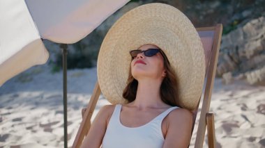 Sahilde eğlenen mutlu bayan, dinlenip hasır şapkayla güneşleniyor. Güneş gözlüğü takan kaygısız kadın şemsiyenin altında kumlu sahilde dinleniyor. Rahatlamış esmer, tatilini okyanus kıyısında geçiriyor..