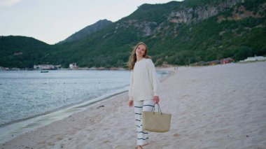 Kum kıyısında çanta taşıyan bir gezgin. Kumsaldaki sakin sahil atmosferinin keyfini çıkaran rahat bir kadın yeşil tepelerin etrafını sarmıştı. Çizgili pantolonlu ve rahat kazaklı gülümseyen bayan boş deniz kıyısına basıyor.