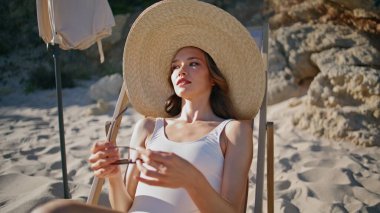 Kaygısız bayan kumlu yaz plajında güneşleniyor. Güneş gözlüğü takan hasır şapkalı şık bir model. Güzel deniz kıyısında gün ışığının tadını çıkaran çekici bir kadın gezgin..