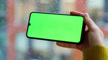 Kromakey akıllı telefon çalan kadın yağmurlu pencerenin yanında bekliyor. Çevrimiçi alımları yeşil ekranlı cep telefonundan seçen kimliği belirsiz bir kadın. Kadın elleri yatay model cep telefonlarını tutuyor..