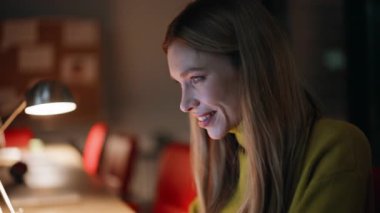 Karanlık işyerinde ışık altında fazla mesai yapan gülümseyen bir çalışan. Gece ofis alanında dizüstü bilgisayarını okuyan mutlu bir iş kadını. Çalışkan kadın, gece geç saatlerde kapalı alanda proje geliştiriyor..