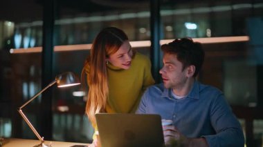 Gülümseyen meslektaşlar akşam geç saatlerde dizüstü bilgisayarın açılışını tartışıyorlar. Gece ofisindeki projede birlikte çalışan mutlu iki yönetici. Neşeli iş arkadaşları karanlık kurumsal işyerlerine beyin fırtınası yapıyorlar..