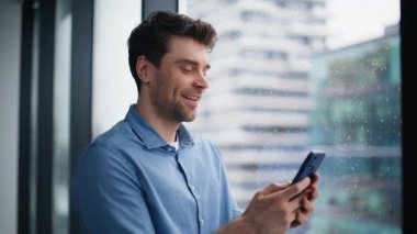 Gülümseyen işadamı ofis panoramik penceresinin yanında cep telefonu mesajı okuyor. Mutlu çalışan maaş bildirimi alıyor. Neşeli menajer cep telefonundan sosyal medyayı izliyor.