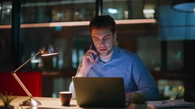 Gece ofis kapanışında birden fazla iş yürüten CEO konuşan cep telefonu görünümlü dizüstü bilgisayar görüntüsü. Gülümseyen işadamı internet sitesini arıyor. Mutlu adam yönetici karanlık iş yerinde akıllı telefon konuşuyor..