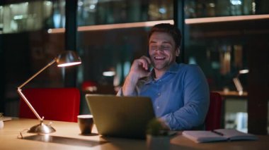 Mutlu profesyonel telefon konuşmaları akşam üzeri ofis kapanışında. Gülümseyen işadamı akıllı telefonu arıyor ve dizüstü bilgisayarın başında oturuyor. Neşeli adam yönetici gece işyerinde cep telefonundan konuşuyor..
