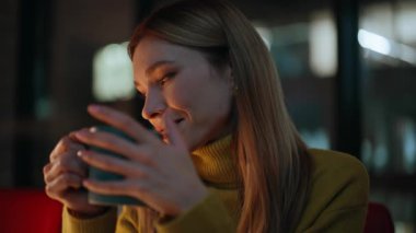 Karanlık dairede elinde kahve fincanı tutan gülümseyen kadın. Portre, esnek sarışın ve bardakta sıcak içecekle evde yalnız oturuyor. Sakin bir iş kadını akşam iş yerinde mesaiye ara veriyor.