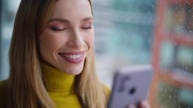 Akıllı telefon sohbeti okuyan neşeli bayan yağmurlu pencerenin yanında dikiliyor. Gülümseyen iş kadını dairede cep telefonuna mesaj atıyor. Evde cep telefonuyla mesajlaşan çekici bir kadın. İletişim konsepti.