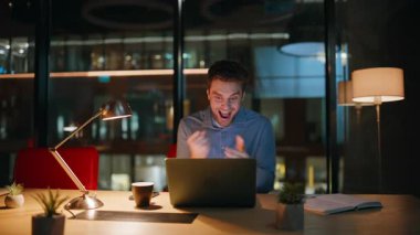 Karanlık işyerinde bilgisayar ekranı izleyerek online zafere sevinen heyecanlı bir gece çalışanı. Coşkulu iş adamı, gece ofisindeki iş başarısının tadını çıkarıyor. Mutlu serbest çalışan adam bilgisayardan harika haberler okuyor..