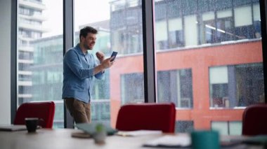 Heyecanlı kazanan ofis odasında akıllı telefon ekranına bakıyor. Duygusal iş adamı pencerenin önünde cep telefonu mesajıyla seviniyor. Kapalı mekanda cep telefonu mesajlarını okumaktan mutlu olan adam.