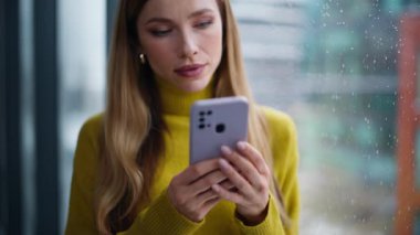 Yağmurlu pencerenin yanında cep telefonuyla mesajlaşan yakın plan kadın. Gülümseyen iş kadını internette sohbet ediyor. Ev telefonuna bakıyor. Güzel bir bayan cep telefonu uygulaması kullanarak internet alışverişini seviyor..