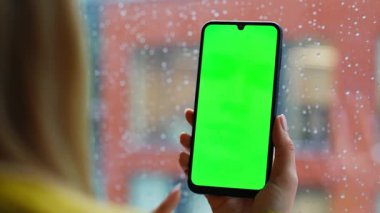 Kadın elleri yeşil ekran cep telefonu araklıyor. İnternetten ev alışverişi yapıyor. Bilinmeyen iş kadını, yağmurlu bir pencerede duran yeşil ekran akıllı telefondan mesaj arıyor. Model telefon kullanan kadın.