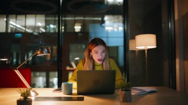 Başarılı bir projeye tepki gösteren sürpriz iş kadını duygusal olarak gece ofisinde oturuyor. Karanlık işyerinde dizüstü bilgisayar arayan aşırı neşeli bir kadın. Gülümseyen bayan heyecanlanıyor, el çırpıyor..