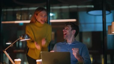 Heyecanlı ortaklar, karanlık ofiste dizüstü bilgisayar görüntüsü alarak seviniyor. Mutlu iki meslektaş gece işyerinde iş başarısını kutluyor. Duygusal iş arkadaşları beş kişiye başarıyı tattırıyor.