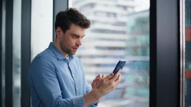 Akıllı telefonun sevinç çığlıklarını izleyen gülen adam yakın plan kazandı. Heyecanlı iş adamı ofis penceresinin önünde cep telefonundan harika haberler okuyor. Çok mutlu bir girişimci, duygusal bir şekilde telefonu elinde tutuyor.