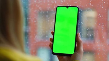 İşkadını parmağı kaydırmalı cep telefonu yağmurlu pencerenin önünde duruyor. Tanımlanamayan bir kadın evdeki yeşil ekran cep telefonundan haberleri izliyor. Bayan tarayıcı uygulaması Chroma anahtar akıllı telefon.
