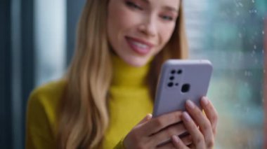 Kadın eli yakın çekimde akıllı telefondan mesaj yolluyor. Gülümseyen güzel bayan pencerenin önünde durup sosyal medyayı izliyor. Cep telefonuyla mesajlaşan mutlu sarışın portresi. İletişim konsepti