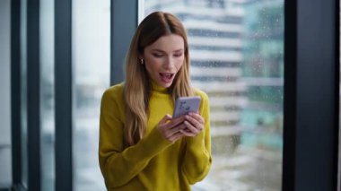 Cep telefonundan mesaj alan heyecanlı kadın duygusal olarak yakın çekim yapıyor. Akıllı telefondan harika haberler okuyan mutlu serbest çalışan kadın başarılı oldu. Pencerenin kenarında bekleyen çok mutlu bir bayan..