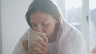 Sakin bir kadın, sakin bir sabahta kahvesini yudumluyor. Battaniyeli gülümseyen kız gün ışığında sıcak içeceğin tadını çıkarıyor. Güzel bayan huzur içinde tek başına dinleniyor. Hasta kadın bitki çayını yudumluyor.