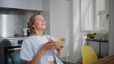 Neşeli kız güneş ışığında mısır gevreği yiyor. Masada kahvaltıyı karıştıran mutlu kadın eldiveni. Pijamalı gülümseyen kadın rahat ev atmosferinin tadını çıkarıyor. Neşeli bayan sabah istirahat ediyor..