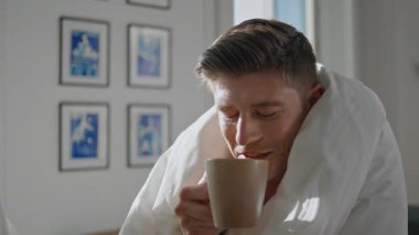 Adam battaniyeye sarılı ev kahvesi içiyor. Portre, sabah güneşinde sıcak bir içecek içip bilinmeyen partnerle konuşmaktan zevk alıyor. Sıcak atmosferde kahvaltı yapan gülümseyen yakışıklı erkek. Ev eğlencesi yaşam tarzı