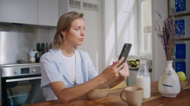 Evde akıllı telefon kullanan rahat bir kadın. Kapalı çekim sürpriz kız hafta sonu kahvaltı yaparken sosyal medyayı kontrol ediyor. Gülümseyen genç bayan cep telefonuyla sabah rutininin keyfini çıkarıyor.