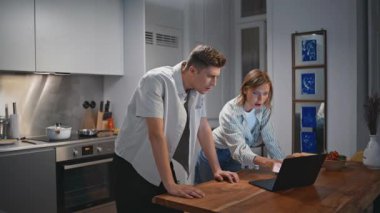Genç eşler evde birlikte yemek pişiriyor. Modern mutfakta laptoptan yemek videosu izleyen neşeli insanlar. Gülümseyen kadın akşam yemeği hazırlayan kocadan bahsediyor. Mutlu aşıklar çevrimiçi tarif arıyor