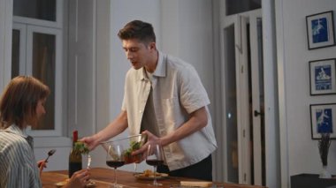 Gülümseyen koca modern yemek odasında yemek servisi yapıyor. Neşeli koca konuşan eş evde romantik bir akşam geçiriyor. Mutlu evli çift hafta sonu ilişki bağlarını kutluyor. Boş zaman yaşam tarzı.