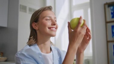 Güneşli mutfakta lezzetli elmaları ısıran bir kadın. Kapanış kızı taze meyvelere hayran evde huzurlu bir sabah geçiriyor. Sağlıklı atıştırmalıklar yiyen gülümseyen kadın içeride kameraya bakıyor. Refah diyeti konsepti.