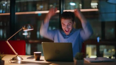 Mutlu kazanan dizüstü bilgisayara bakıyor ve gece ofisindeki iş başarısını kutluyor. Heyecanlı iş adamı projenin başarısından duygusal olarak zevk alıyor. Büyük sevinçle bilgisayar başında harika haberler okuyor..