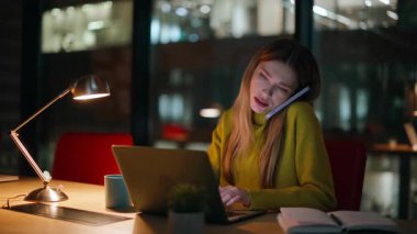 Fazla mesai çalışanı cep telefonuyla konuşuyor karanlık ofiste oturuyor. Esneyen iş kadını, gece iş yerinde akıllı telefondan arıyor bilgisayarını arıyor. Yorgun kadın serbest çalışan motivasyonsuz endişeli son teslim tarihi