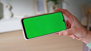 Elinde yeşil ekran akıllı telefon olan bir kadın evde video izliyor. Bilinmeyen kız yatay model cep telefonunda sosyal medya içeriğini araştırıyor. Kadın serbest çalışan Chroma Key telefonunda webinar görünümlü.