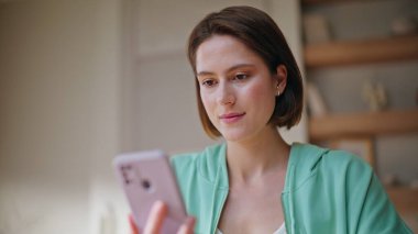 Rahat esmer görünümlü cep telefonu ekranı evde yakın çekimde internet iletişiminin keyfini çıkarıyor. Oturduğu dairede akıllı telefon mesajı okuyan gülümseyen genç kadın. Telefon internet uygulamasında pozitif kadın alışverişi.