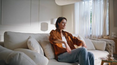 Kaygısız kadın yumuşak beyaz yastıklarla kanepeye düşüyor. Gülümseyen kadın rahat bir kanepede oturur ve hafta sonu evde tek başına dinlenir. Mutlu kadın esmer modern dairede rahat dinleniyor.