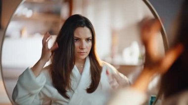Güzel bir bayan yatak odasındaki aynada saçına dokunuyor. Beyaz cüppeli rahat kadın saç stilini ayarlıyor. Sakin, uzun saçlı esmer kadın evde günlük saç bakımını yapıyor.