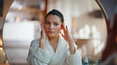 Esmer, ayna yansımasının pürüzsüz cilde yakından dokunmasından hoşlanır. Beyaz cüppeli gülümseyen kadın cilt bakımına hayran. Mutlu bayan günlük bakım prosedüründen memnun..