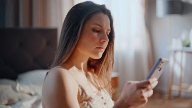 Erken saatte cep telefonuna mesaj atan, güneşli bir sabah yatağında oturan bir kadın. Dairenin yatak odasında esmer, akıllı telefon mesajları. Modern bir dairede uyandıktan sonra SMS yazan çekici bir kadın..