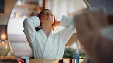 Ev kızı aynaya bakıp beyaz bornoz giyip saç stili yapıyor. Gülümseyen güzel bir kadın at kuyruğu yapıyor ve sabah rutininde sağlıklı saçların tadını çıkarıyor. Mutlu esmer yatak odasındaki yansımaya hayran..