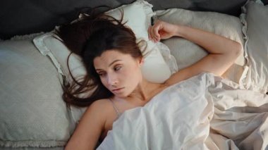 Uyanık bayan, tek başına yumuşak yatağa uzanmış akıllı telefondan saati kontrol ediyor. Üst tarafı yakın çekim. Uykulu bir kadın yatakta cep telefonuna bakarak uyanıyor. Uykusuz esmer, battaniyeye saklanmış, yorgun.