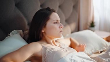 Uykulu kadın baş ağrısı çekiyor. Sıcak bir evde uyanıyor. Yakın plan. Mutsuz kadın, erken uyandıktan sonra yatak odasında baş ağrısı çekiyor. Üzgün genç esmer, sabahları sağlık sorunları yaşıyor..