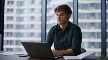 Motivasyonlu yönetici modern ofis alanındaki bilgisayarı daktilo ediyor. Panoramik pencerede kamerada gülümseyen profesyonel bir çalışan. Modern işyerinde şık bir iş adamı pozu.