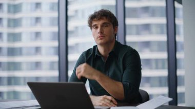 Zengin girişimci ofis çalışma yerinde kameraya güvenle yakın çekim yapıyor. Dizüstü bilgisayarda çalışan yakışıklı iş adamı elinde kalem tutuyor. Modern şirket içinde ciddi bir yönetmen pozu..
