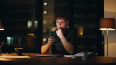 Kapalı dizüstü bilgisayarla karanlık bir iş yerinde cep telefonuna mesaj atan mutlu gece çalışanları. Gülümseyen adam, lamba ışığı altında akıllı telefondan mesaj atıyor. Rahat iş adamı geceleri cep telefonunda e-posta okuyor..