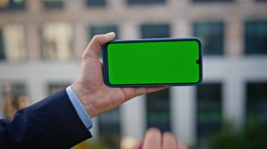 Modern mimari arka planda akıllı telefon uygulaması yakın planda. Bilinmeyen patron yeşil ekran cep telefonunu izliyor. İş adamı şehirde çalışmak için kromakey cep telefonu kullanıyor