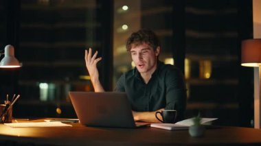 Gülümseyen iş adamı, gece iş yerinde dizüstü bilgisayarla konuşuyor. Karanlık ofiste videoyla projeyi tartışan pozitif serbest yazar. Yakışıklı girişimci gecenin geç saatlerinde sanal toplantıdan zevk alır..