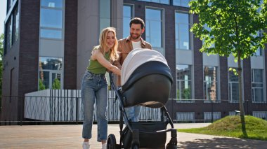 Modern yaşam kompleksinde bebekle yürüyen mutlu bir çift. Sevgi dolu, genç anne, baba ve çocuk güneşli bir günün tadını çıkarıyorlar. Bebek arabasında küçük çocuğu olan gülümseyen bir kadın.