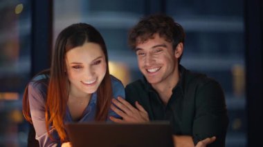 Karanlık ofiste kameraya çekilen iki çalışanın gülümsemesi. Mutlu iş ortakları iş yerinde yapılan görüşmeleri bitirdiler. Pozitif meslektaşlar gece geç saatlerde sanal konferansın keyfini çıkarıyorlar.